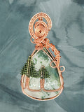 Copper wire wrapped Tree Agate pendant with green wire pine trees and spiral copper bail on blue-gray background